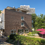 Haus Oldenburg, ein Backsteinhaus mit Balkonen der Ferienanlage Timmendorfer Strand, mit Terrasse und Sonnenschirmen im Vordergrund, Bäume, ein weißes Hochhaus und blauer Himmel im Hintergrund.