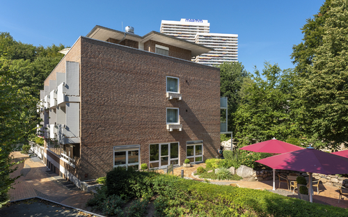 Haus Oldenburg, ein Backsteinhaus mit Balkonen der Ferienanlage Timmendorfer Strand, mit Terrasse und Sonnenschirmen im Vordergrund, Bäume, ein weißes Hochhaus und blauer Himmel im Hintergrund.