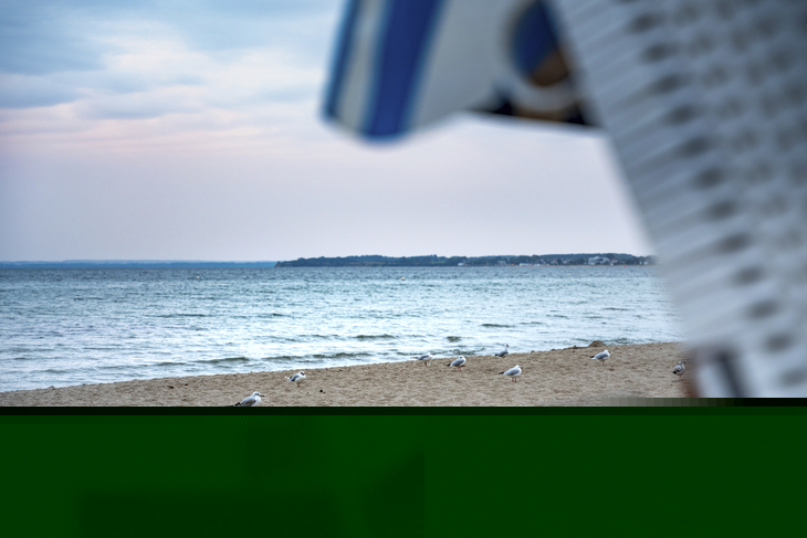 Rechts im Bild ein Teil von einem Strandkorb sowie ein Küstenstreifen der ins Meer ragt. Sandstrand mit Möven, dahinter das Meer bei leichter Dämmerung. 