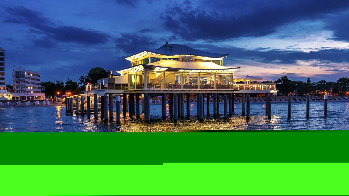 Blick vom Meer auf die Seebrücke Timmendorf mit beleuchtetem Gebäude bei Abenddämmerung, sowie Häusern an der Küste im Hintergrund.