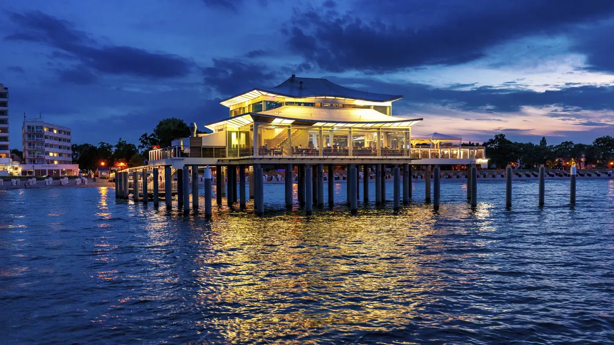 Blick vom Meer auf die Seebrücke Timmendorf mit beleuchtetem Gebäude bei Abenddämmerung, sowie Häusern an der Küste im Hintergrund.