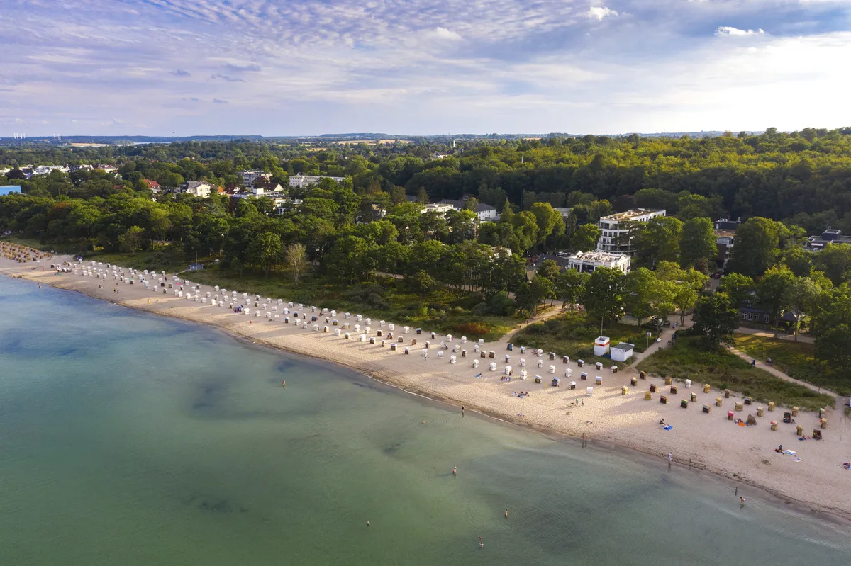 Luftaufnahme von Timmendorfer Strand mit vielen Strandkörben und dahinterliegendem Wald mit Häusern dazwischen, bei leicht bewölktem Himmel.