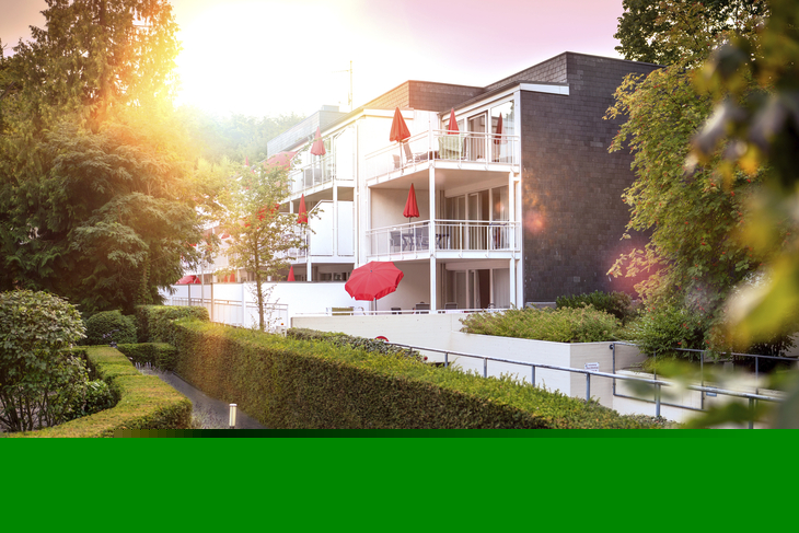 Haus Christel, ein weißes Gebäude der Ferienanlage Timmendorfer Strand, mit Balkonen und roten Sonnenschirmen im Sonnenlicht. Im Vordergrund grüne Hecken und Bäume.
