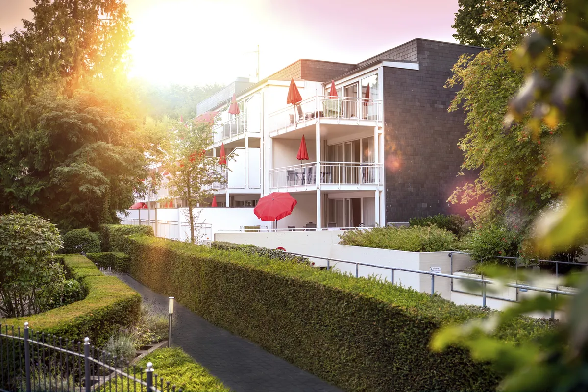 Haus Christel, ein weißes Gebäude der Ferienanlage Timmendorfer Strand, mit Balkonen und roten Sonnenschirmen im Sonnenlicht. Im Vordergrund grüne Hecken und Bäume.