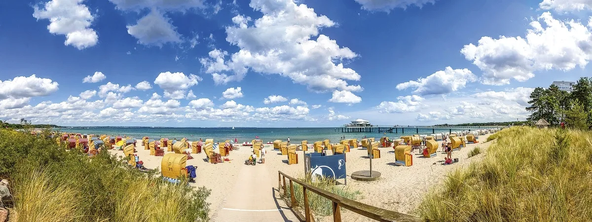 Blick vom Weg zwischen den mit Seegras bewachsenen Dünen auf den Strand mit Strandkörben, dem Meer und blauem Himmel mit Wolken am Timmendorfer Strand.