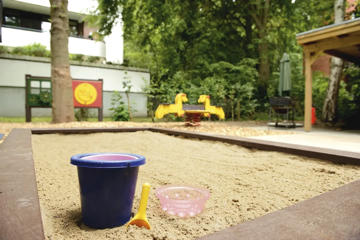 Sandkasten mit Eimer, Schaufel und Förmchen im Vordergrund und Wipptier im Hintergrund, sowie Bäume und die Ecke eines Hauses, in der Ferienanlage Timmendorfer Strand.