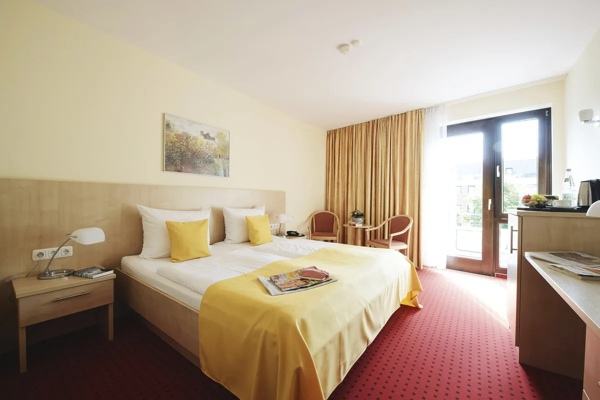 Helles Zimmer mit rotem Teppichboden im Haus Oldenburg in der Ferienanlage Timmendorfer Strand. Doppelbett mit gelber Bettwäsche, Nachtischen und zwei Stühlen mit kleinem Tisch, sowie einem Schreibtisch und Balkontür mit großem gelb-rotem Vorhang.