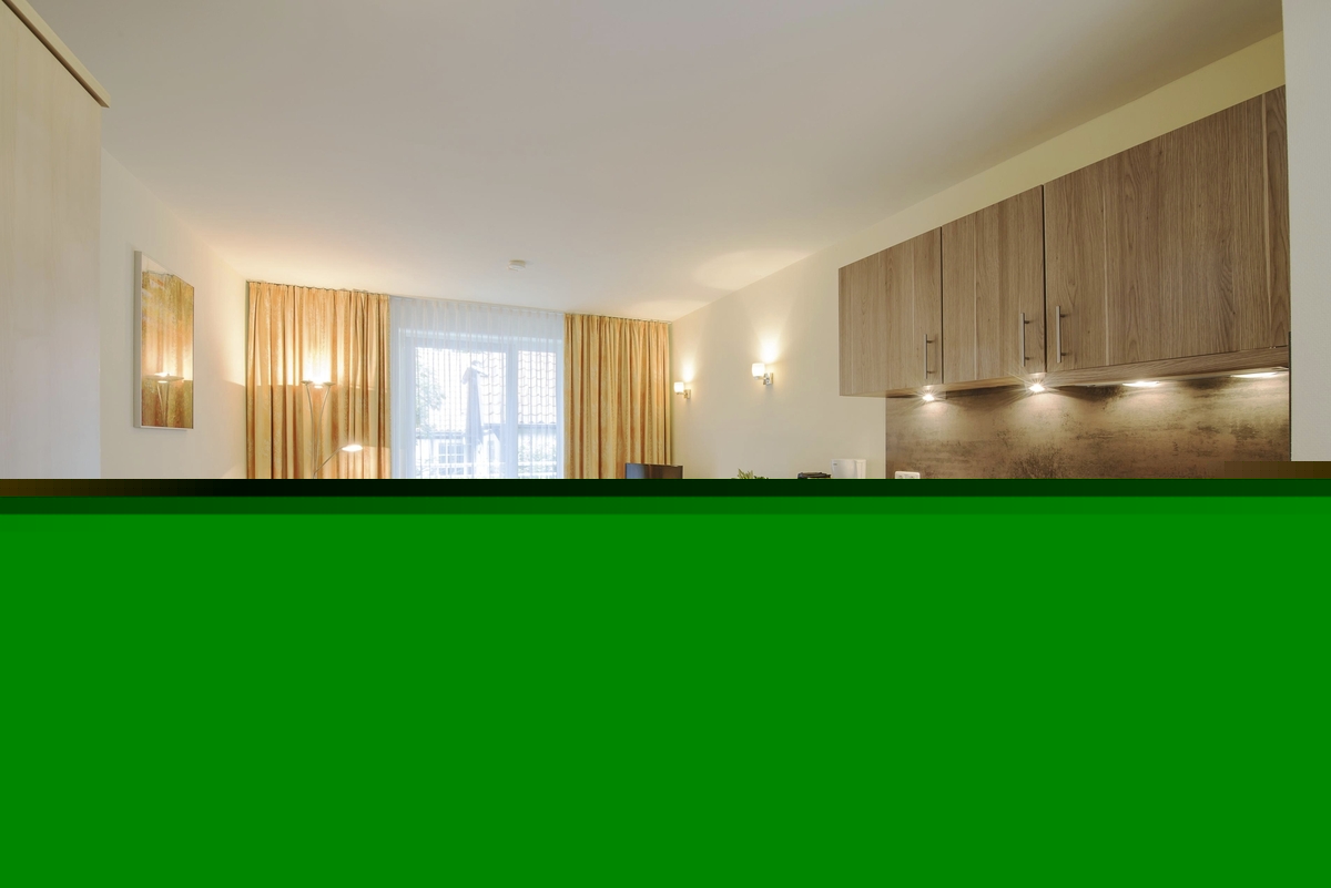 Wohnspiel Haus Oldenburg in der Ferienanlage Timmendorfer Strand. Rechts im Bild eine moderne Küchenzeile, links blaues Sofa und zwei Sessel mit kleinem runden Tisch. Hinten im Bild ein großes Fenster mit Vorhängen rechts und links und einem Fernseher davor.