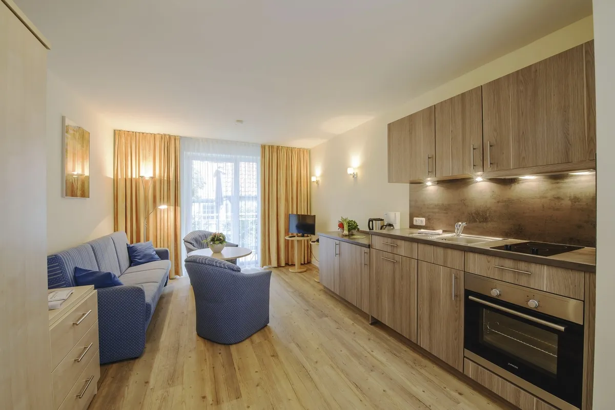 Wohnspiel Haus Oldenburg in der Ferienanlage Timmendorfer Strand. Rechts im Bild eine moderne Küchenzeile, links blaues Sofa und zwei Sessel mit kleinem runden Tisch. Hinten im Bild ein großes Fenster mit Vorhängen rechts und links und einem Fernseher davor.