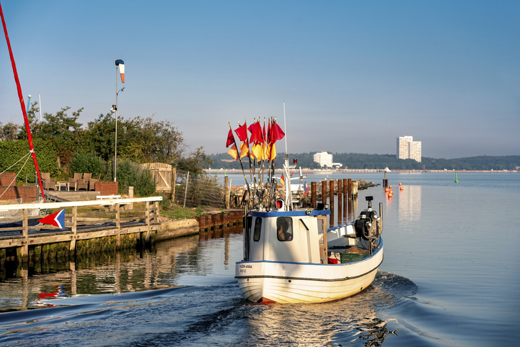 Ein kleines Fischerboot im Hafen von Niendorf bei klarem Himmel, weißes Hochhaus umgeben von Wald im Hintergrund.