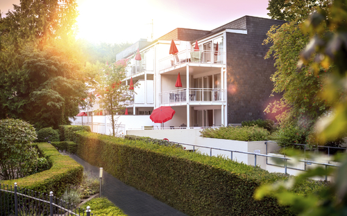 Haus Christel, ein weißes Gebäude der Ferienanlage Timmendorfer Strand, mit Balkonen und roten Sonnenschirmen im Sonnenlicht. Im Vordergrund grüne Hecken und Bäume.