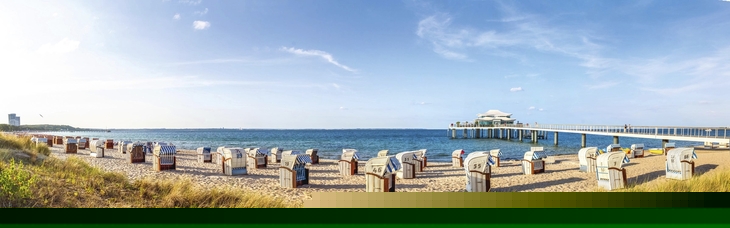 Strandkörbe am Timmendorfer Strand mit Seebrücke im Hintergrund und Seegras im Vordergrund, bei leichtbewölktem Himmel.