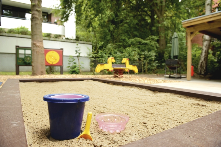 Sandkasten mit Eimer, Schaufel und Förmchen im Vordergrund und Wipptier im Hintergrund, sowie Bäume und die Ecke eines Hauses, in der Ferienanlage in Timmendorfer Strand.