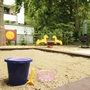 Sandkasten mit Eimer, Schaufel und Förmchen im Vordergrund und Wipptier im Hintergrund, sowie Bäume und die Ecke eines Hauses, in der Ferienanlage in Timmendorfer Strand.