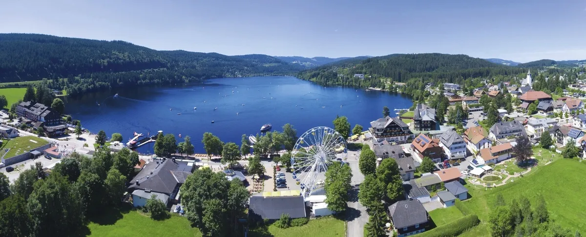 Luftaufnahme eines Sees mit Riesenrad und umliegender Stadt in einer grünen Landschaft.