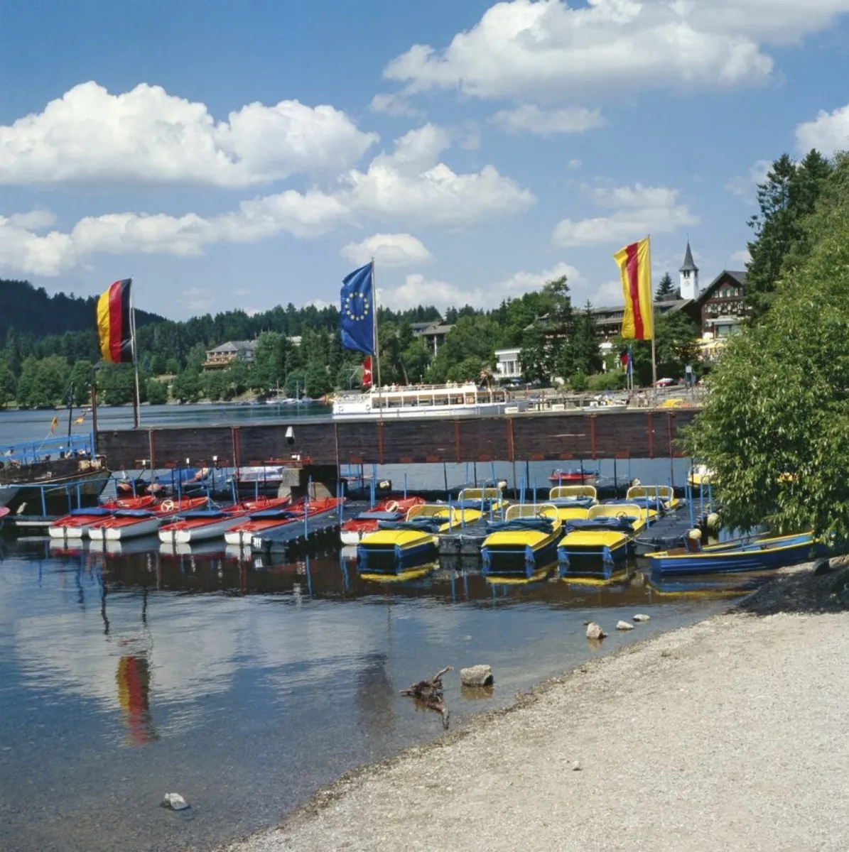 Boote an einem Steg am Titisee, umgeben von Bäumen und einer Ferienanlage im Hintergrund.