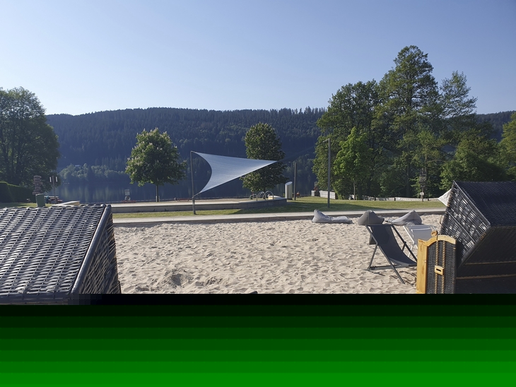 Sonnenliegestühle und Strandkörbe an einem Sandstrand mit Blick auf den Titisee und Wald.