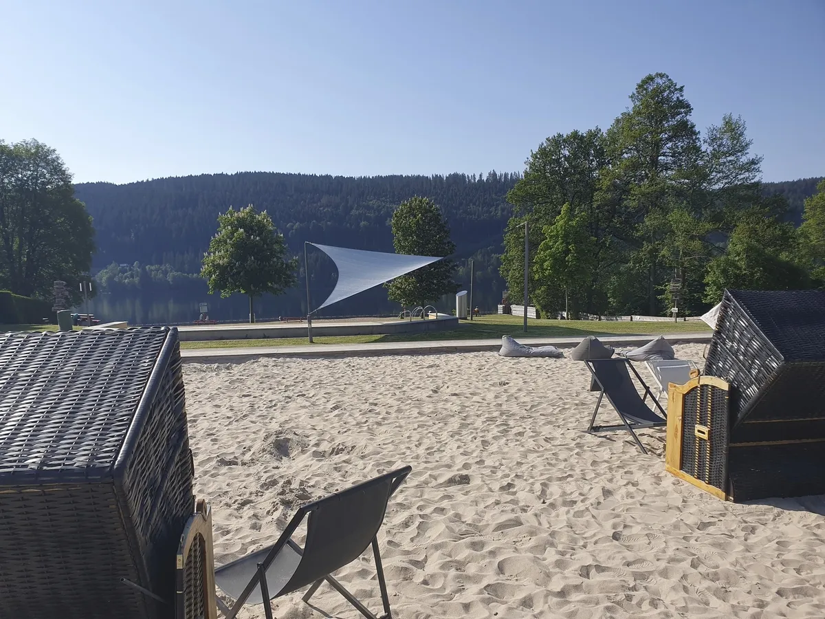 Sonnenliegestühle und Strandkörbe an einem Sandstrand mit Blick auf den Titisee und Wald.