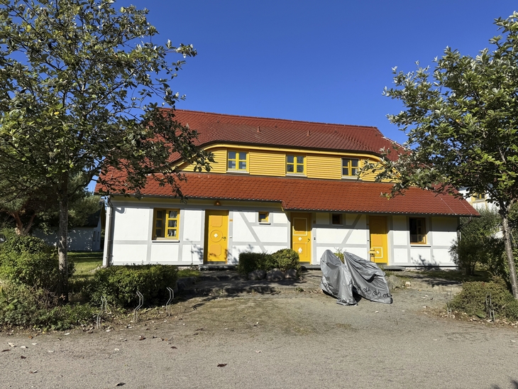 Ferienhaus mit gelber Tür im Feriendorf, umgeben von Bäumen bei sonnigem Wetter.