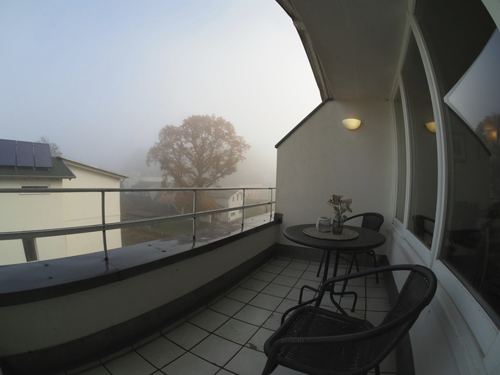 Balkon mit Tisch und Stühlen, Blick auf neblige Landschaft und Baum im Hintergrund.