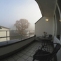 Balkon mit Tisch und Stühlen, Blick auf neblige Landschaft und Baum im Hintergrund.