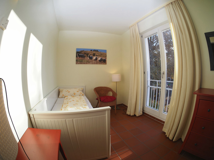 Helles Zimmer mit Bett, Sessel, Lampe und großem Fenster mit Vorhängen.