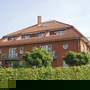 Rotes Backsteingebäude mit Balkon und rotem Dach, umgeben von grüner Hecke.