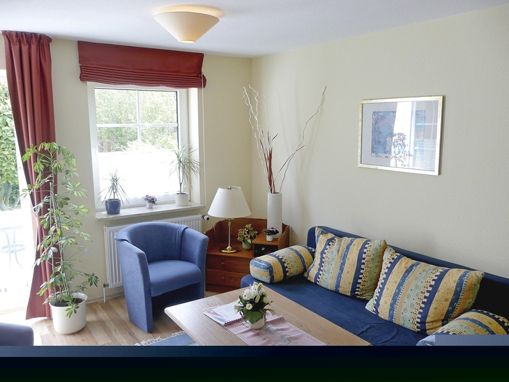 Wohnzimmer mit Sofa, Sessel, Tisch, Pflanzen und einem Fenster mit Vorhängen.