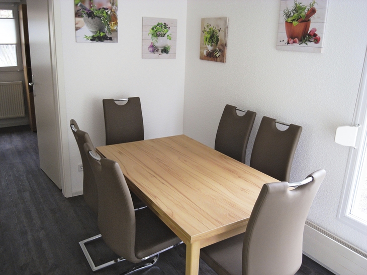 Essbereich mit Holztisch und sechs braunen Stühlen vor weißer Wand mit Bildern.