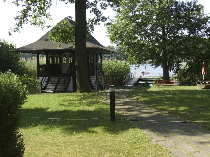 Blick auf ein Teehaus an einem See mit Steg und Bäumen im Vordergrund.