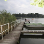 Steg am See mit Booten und Schilf, bewölkter Himmel im Hintergrund.