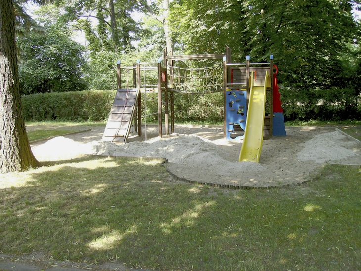 Spielplatz mit Klettergerüst und Rutsche, umgeben von Bäumen und Rasen.