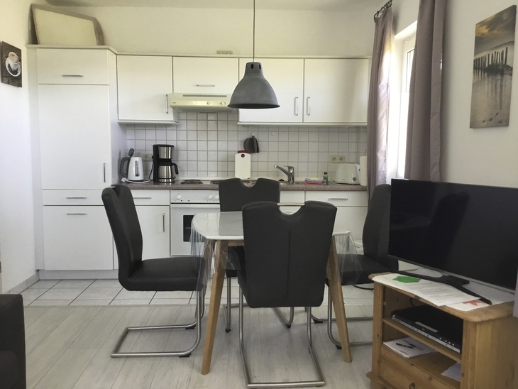 Kleine Küche mit Esstisch, Stühlen, weißen Schränken und Fernseher.