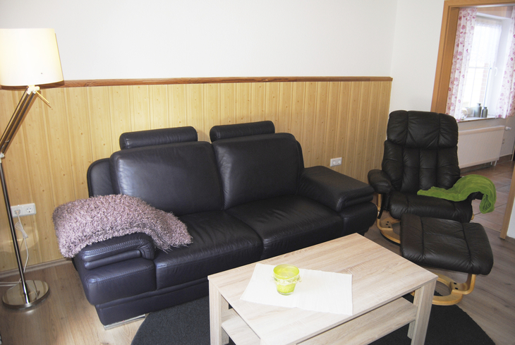 Wohnzimmer mit schwarzem Ledersofa, Sessel und Holztisch, helle Wandvertäfelung.