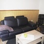 Wohnzimmer mit schwarzem Ledersofa, Sessel und Holztisch, helle Wandvertäfelung.
