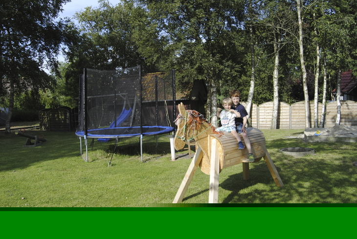 Kinder spielen auf einem Holzpferd neben einem Trampolin auf einem Spielplatz.