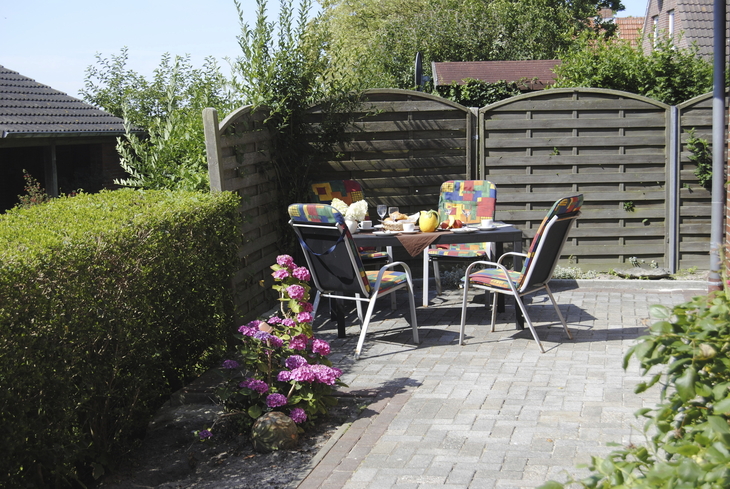 Gartenterrasse mit Tisch, Stühlen und blühenden Blumen im Sonnenschein.
