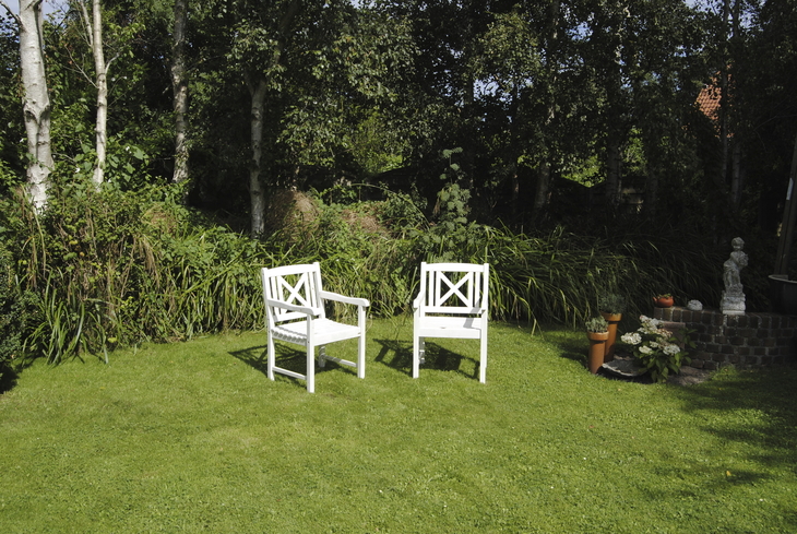 Zwei weiße Gartenstühle auf Rasenfläche, umgeben von Bäumen und Pflanzen.