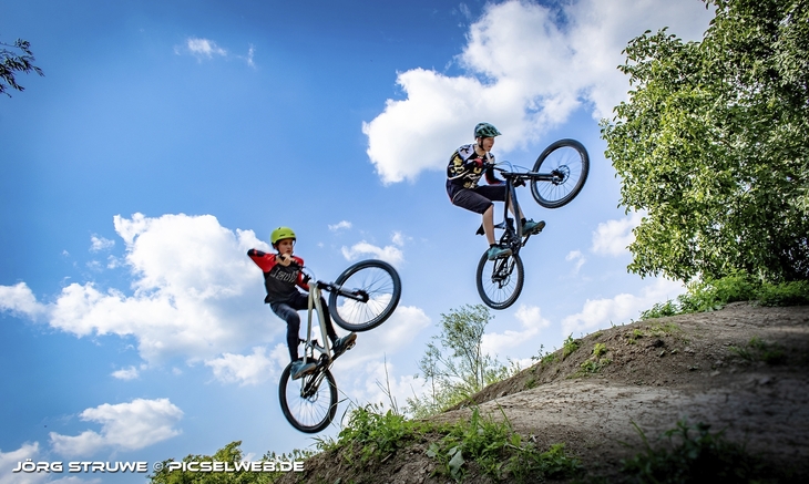 Zwei Personen springen mit Mountainbikes über eine Erhebung im Freien.