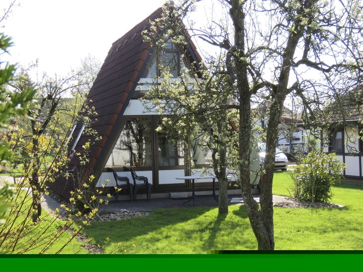 A-förmiges Ferienhaus in sonnigem, grünem Garten mit Bäumen.