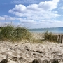 Sandstrand mit Grasbüscheln und Blick auf das Meer unter wolkigem Himmel.