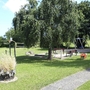 Spielplatz mit Schaukeln und Wippe auf grüner Wiese unter Bäumen.