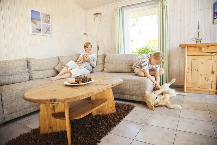 Wohnzimmer mit Sofa, Tisch und zwei Personen, ein Kind spielt mit einem Hund.