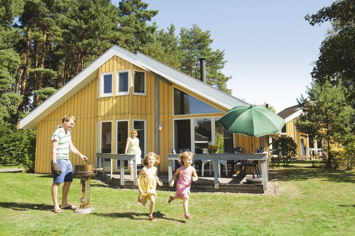 Familie vor einem gelben Ferienhaus im Grünen an einem sonnigen Tag.