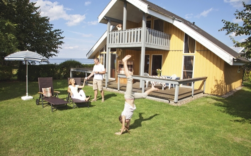 Ein Ferienhaus mit Terrasse und Personen im Garten bei sonnigem Wetter.