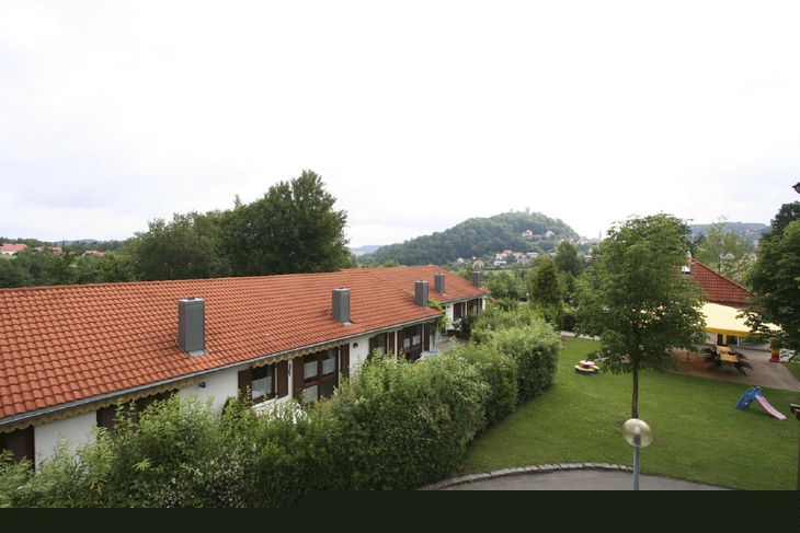 Gebäudekomplex mit roten Dächern und grüner Umgebung, Hügel im Hintergrund.