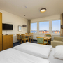 Helles Hotelzimmer mit Doppelbett, Esstisch, Sofa und Fenster mit Blick auf Yachthafen.