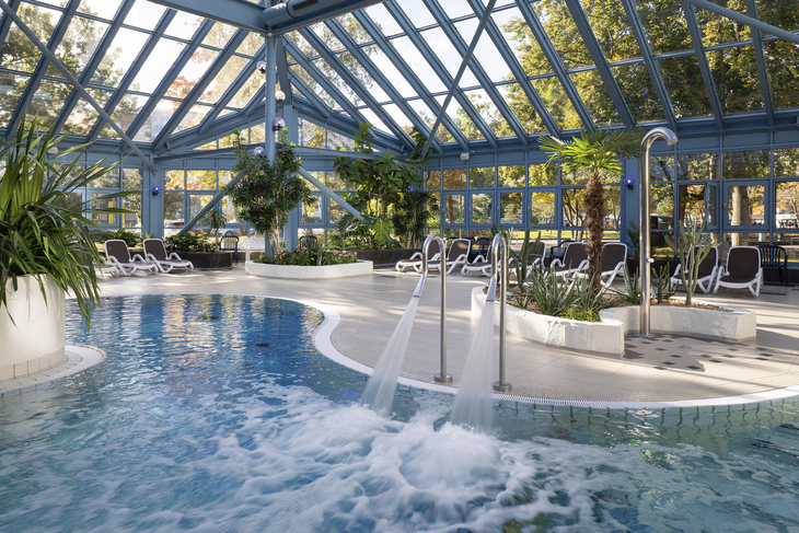 Innenbereich eines Pools mit Glasdach und Liegestühlen im Hintergrund.