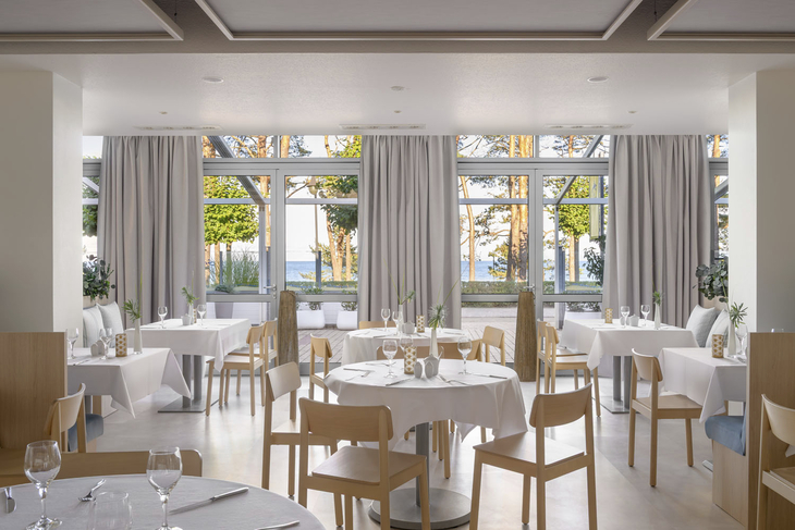 Helles Restaurant mit weißen Tischdecken und Blick auf Bäume und Meer durch große Fenster.