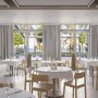 Helles Restaurant mit weißen Tischdecken und Blick auf Bäume und Meer durch große Fenster.